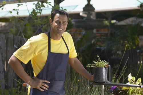 A smiling man wearing a yellow T-shirt and a dark apron stands in a lush garden with diverse plants and greenery, holding a small potted plant with young green shoots. The garden features a variety of foliage, including tall grass-like plants and flowering shrubs, with a paved pathway or decking visible nearby. In the background, a wooden fence and a garden pond or fountain can be seen, along with gardening tools and outdoor furniture partially visible under the shade of trees and a greenhouse, indicating a well-maintained outdoor space suitable for gardening services in Brockley. The scene is illuminated by natural daylight, suggesting a clear weather day, with vibrant colors and healthy plant growth reflecting attentive gardening and outdoor maintenance efforts typical of local garden care providers such as Gardening Services Brockley.